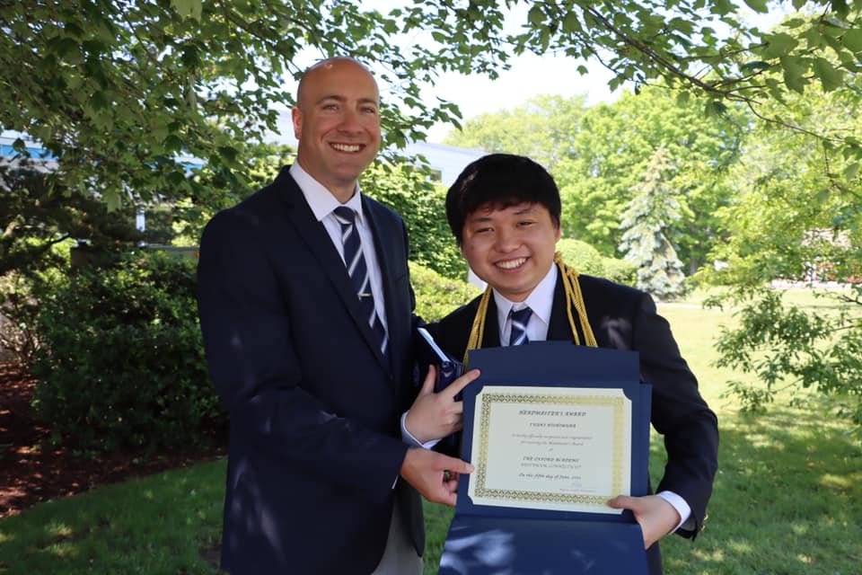 At the end of average, a man in a suit proudly holds a certificate in front of a tree.
