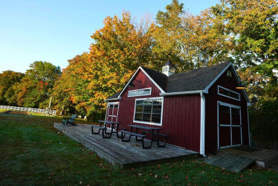 A small red building with white trim, surrounded by autumn trees, features a wooden deck with picnic tables. The sky is clear.