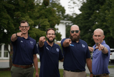 Four men in matching blue shirts, pointing towards the camera, standing outdoors with trees and grass in the background.