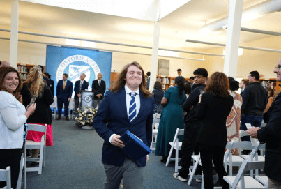 A young man in a school uniform smiling as he walks through a crowd at a graduation ceremony, with people clapping and a banner reading "oxford academy" in the background.