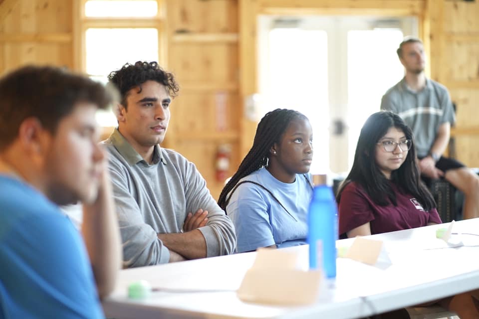 A group of young adults sits at a table in a wooden room, attentively listening—a workshop on executive functioning skills perhaps. One man stands in the background. Blank name cards and a blue water bottle are on the table.