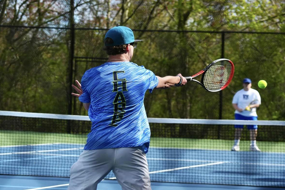 A tennis player in a blue and green shirt hits a ball with a forehand swing while another player in the background observes on an outdoor court at the Oxford Academy in Westbrook, known for its individualized learning approach.
