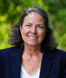 A smiling person with curly hair wearing a dark blazer over a white top stands outdoors with greenery in the background.