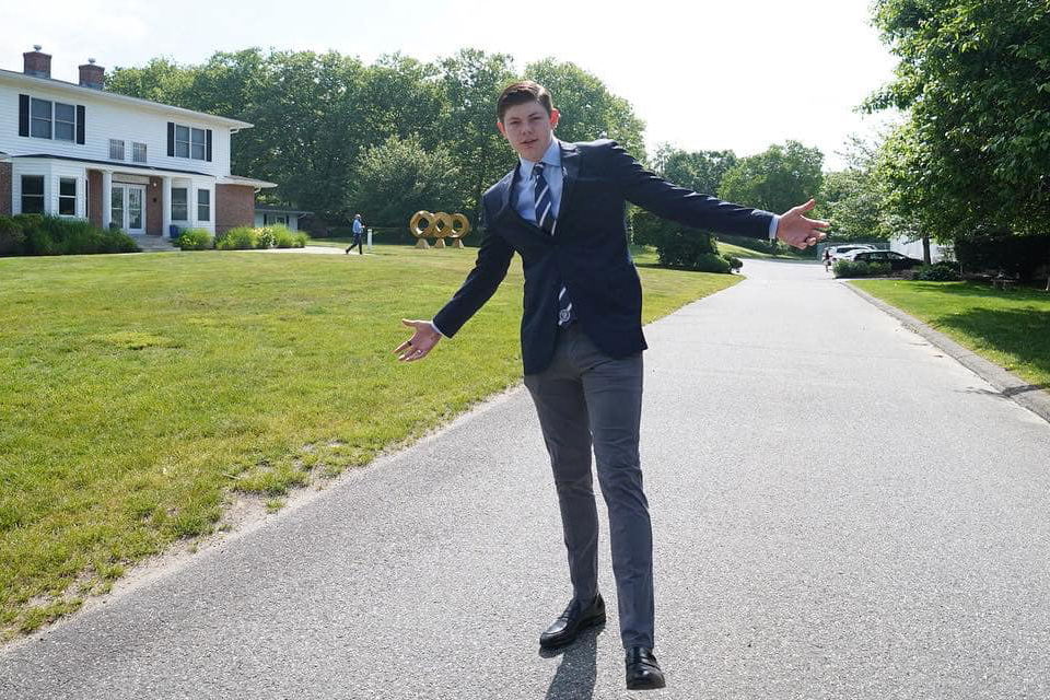 A man in a suit stands on a residential street with arms outstretched, embodying the essence of executive functioning skills. Meanwhile, a person walks in the background near a house and grass.