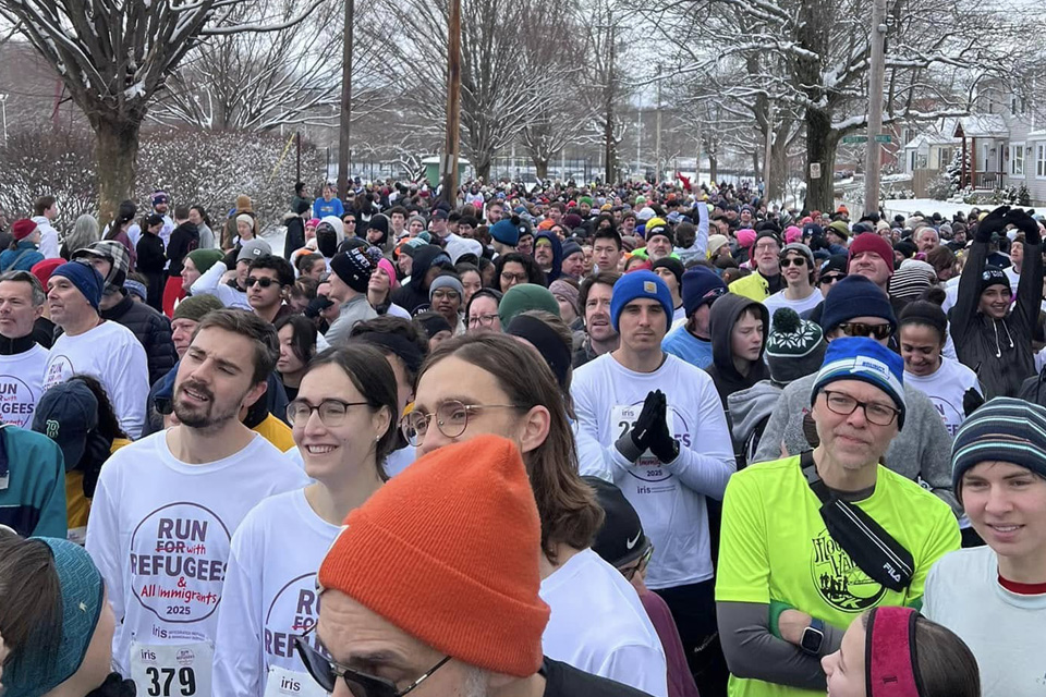 A large group of people, mostly in winter clothing, gather outdoors in Connecticut for the "Run for Refugees" event. Some wear event shirts. Trees and snow are visible in the background, creating a serene atmosphere for this meaningful endeavor.