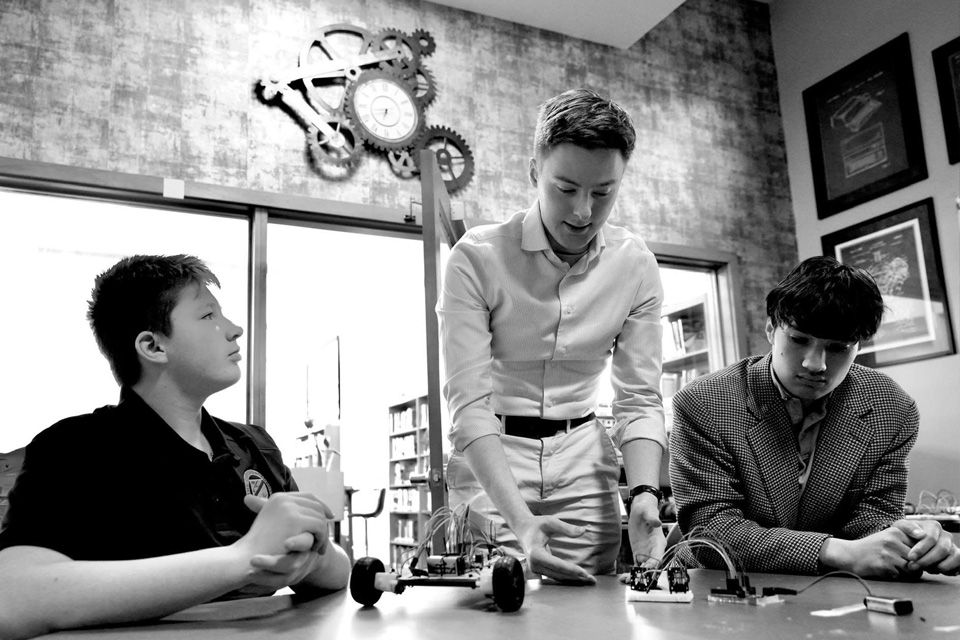 In a vibrant summer session, three people collaborate on small wheeled devices at a table. One stands, explaining to the others, who listen intently. Gears and framed items fill the background, enhancing the focus on improving executive function skills.
