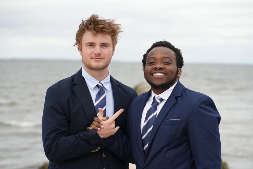Two men in suits and ties stand together by the water in Connecticut, smiling at the camera, with one man making a hand gesture during a Summer Session.