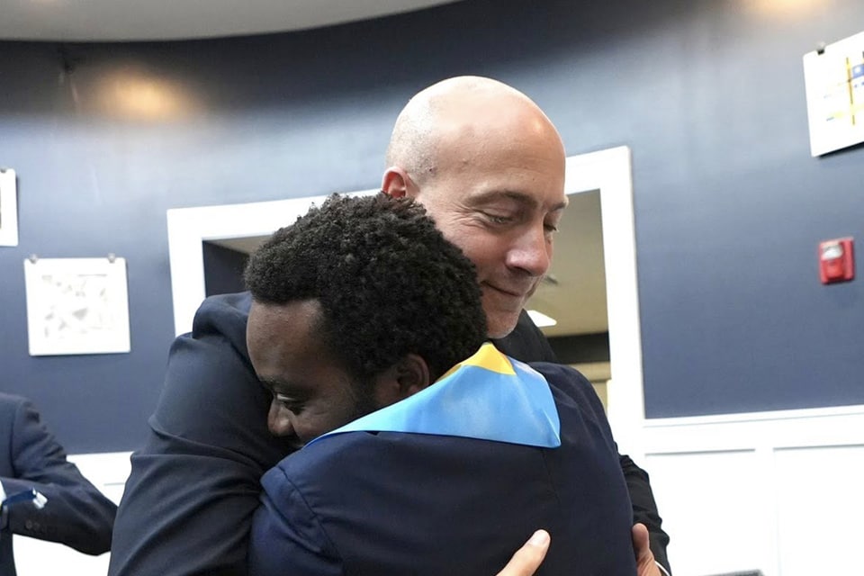 Two people are hugging, one in a blue graduation gown with a yellow stole.