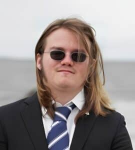 A person with long hair, wearing sunglasses, a dark suit, white shirt, and striped tie, stands outdoors in Connecticut with a blurred gray background.