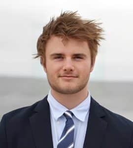 A young man with tousled hair wearing a dark suit, white shirt, and striped tie, standing outdoors against a blurred background, exuding confidence and strong executive function.
