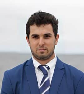 A man in a blue suit and striped tie stands outdoors, looking at the camera with a neutral expression. The blurred background suggests he might be attending a Summer Session focused on individualized learning.