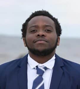 A man in a blue suit and striped tie stands outdoors in connecticut, looking directly at the camera with a neutral expression. The blurred background suggests an overcast sky during a Summer Session.