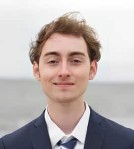 Young man with light brown hair wearing a navy suit and tie, standing outdoors with a neutral background and slight smile—exuding confidence often seen in those thriving through individualized learning or strong Executive Function skills.