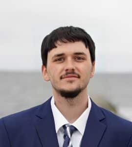 A man in a navy suit and striped tie stands outdoors with a blurred ocean background, embodying executive function and professionalism.