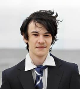 A young man with tousled dark hair wearing a black suit, white shirt, and striped tie stands outdoors in Connecticut.