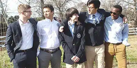 Five young men wearing school uniforms and blazers stand together outdoors, smiling and talking with arms around each other.