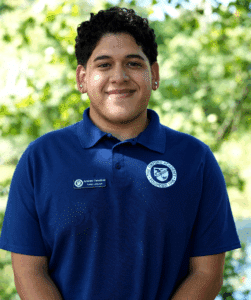 Andres Cevallos, a person with short curly hair and earrings wearing a blue collared shirt and name tag, stands outdoors with greenery in the background.