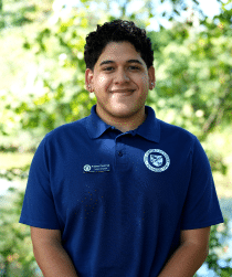 A person named Andres Cevallos, wearing a blue polo shirt with a name tag, stands outdoors in front of green foliage.