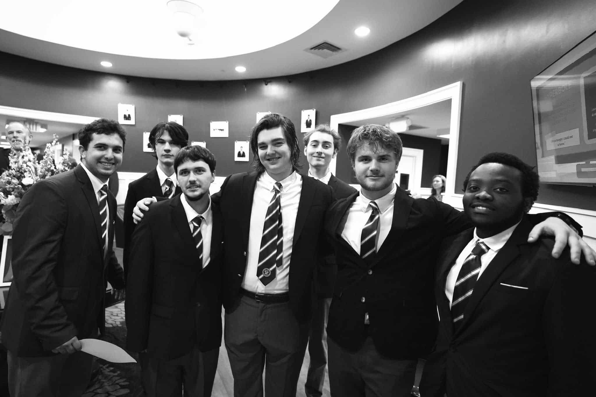 A group of seven young men in suits and striped ties pose together indoors, smiling at the camera, showcasing their bond formed through individualized learning in Connecticut.