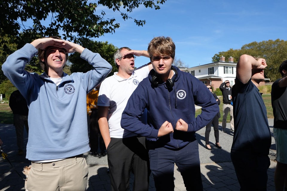 A group of people stand outdoors in daylight, some shielding their eyes from the sun while one person in the center flexes their arms and looks at the camera. A house and trees are in the background, creating a scene fit for a parent guide to outdoor fun.