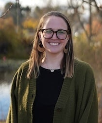 A woman with long hair and glasses, wearing a green cardigan and black top, stands outdoors in front of blurred trees and a pond.