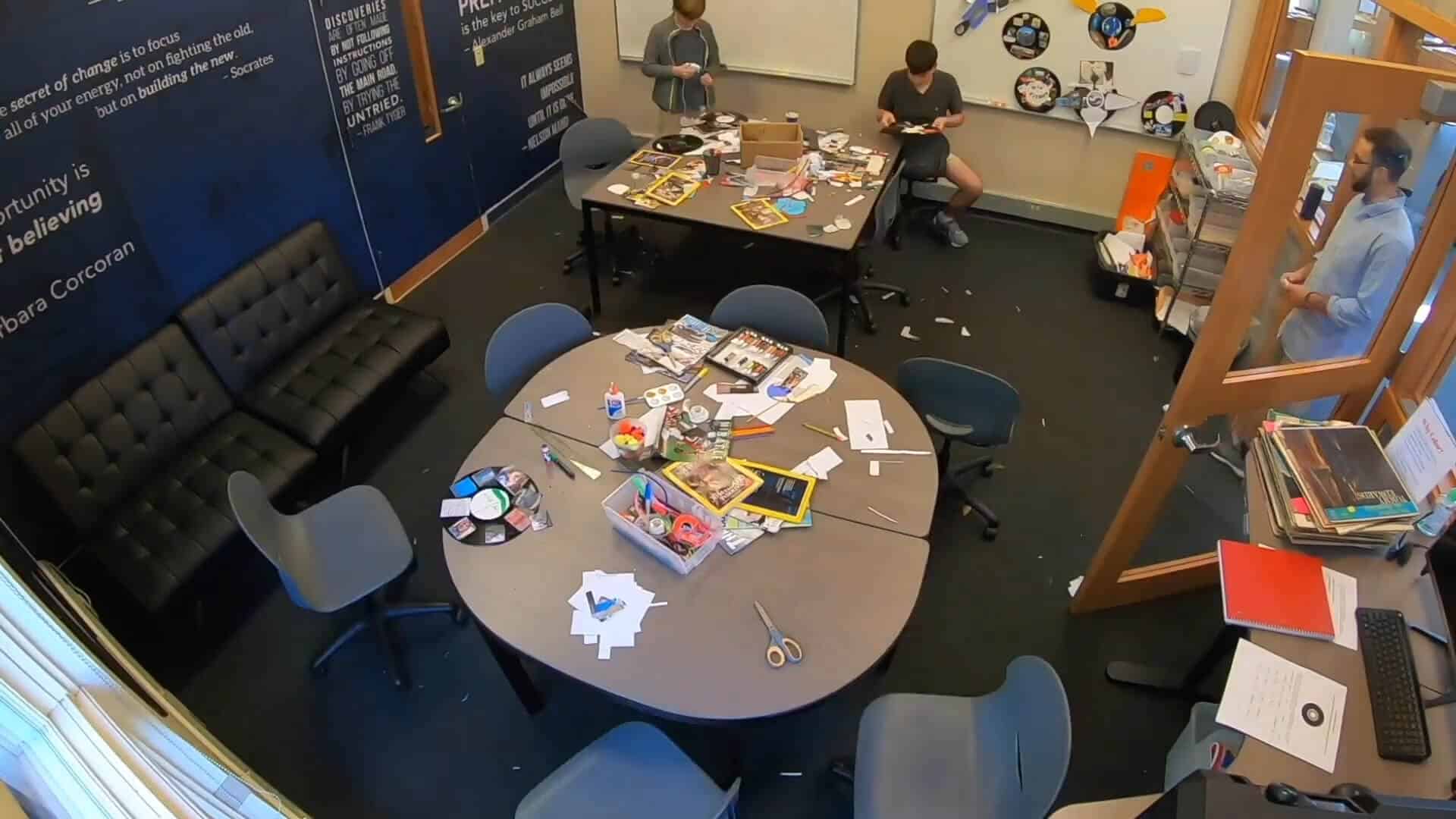 Three people are working and sitting in a classroom or makerspace with tables covered in art supplies, paper, and craft materials, enjoying a creative Summer Experience together.