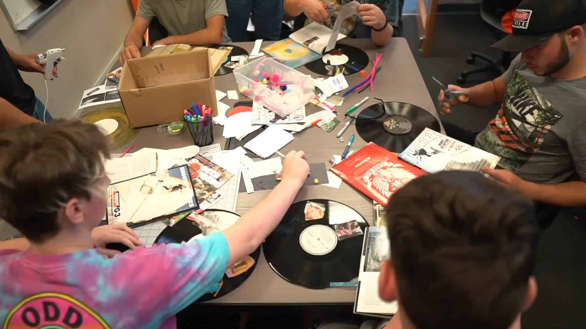 Several people sit around a table covered with vinyl records, craft supplies, and paper, enjoying a collaborative Summer Experience of creative projects together.