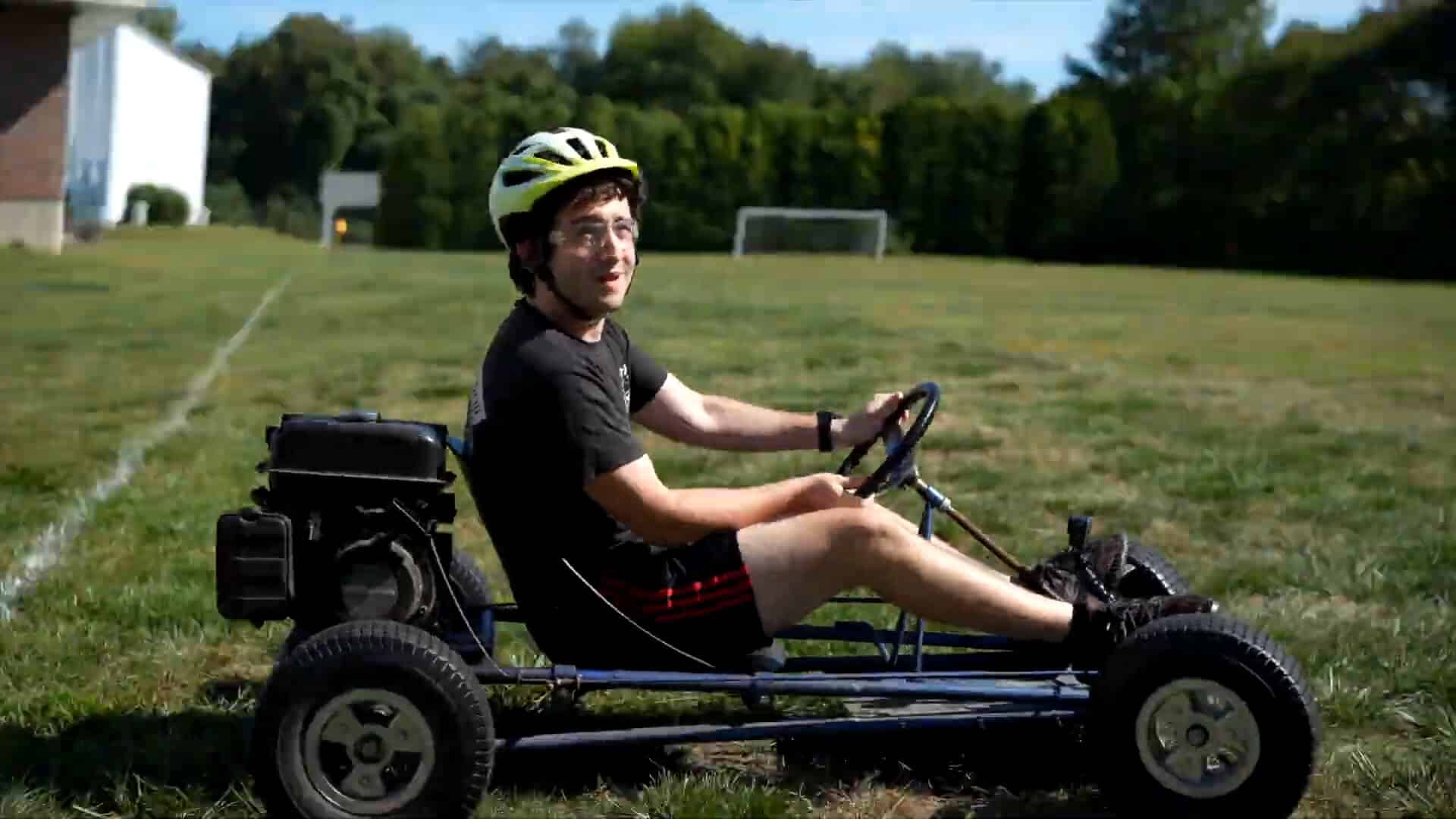 A person wearing a helmet and black athletic clothing sits in a small, open-frame go-kart on a grassy field with trees and a soccer goal—capturing the thrill of an unforgettable Summer Experience.
