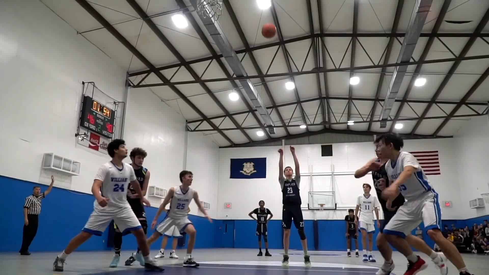 A basketball player in a dark uniform takes a free throw during an exciting Summer Experience as players from both teams and a referee watch in a lively gymnasium.