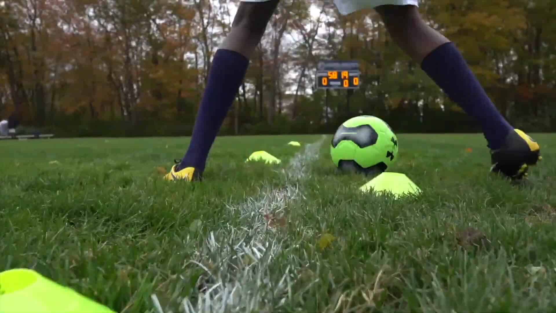 A soccer player stands over a ball placed on a cone during a Summer Experience, on a grassy field with cones and a scoreboard in the background.