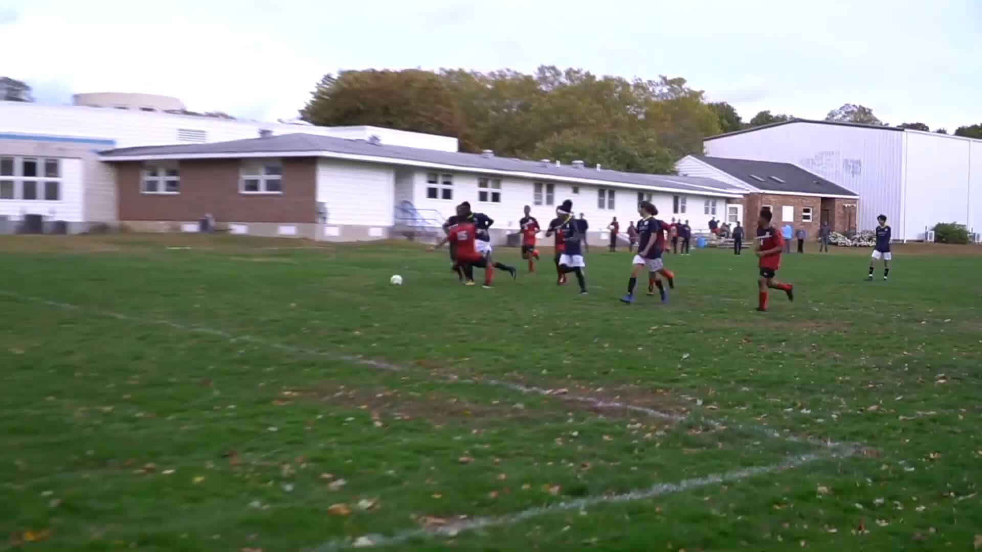 A group of youth soccer players, enjoying a vibrant Summer Experience, run toward a soccer ball on a grassy field in front of school buildings.