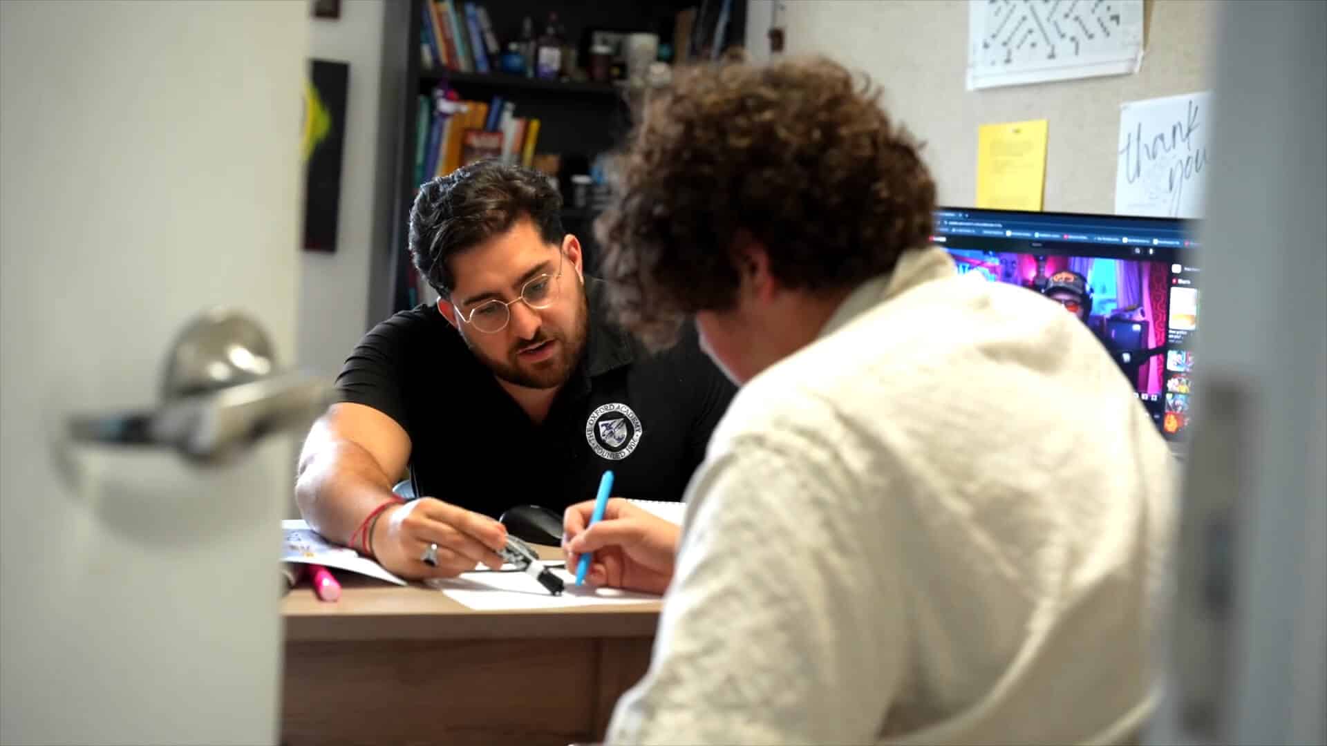 Two people sit at a desk discussing paperwork, one pointing at a document with a pen while the other takes notes about their summer experience, with bookshelves and a computer in the background.