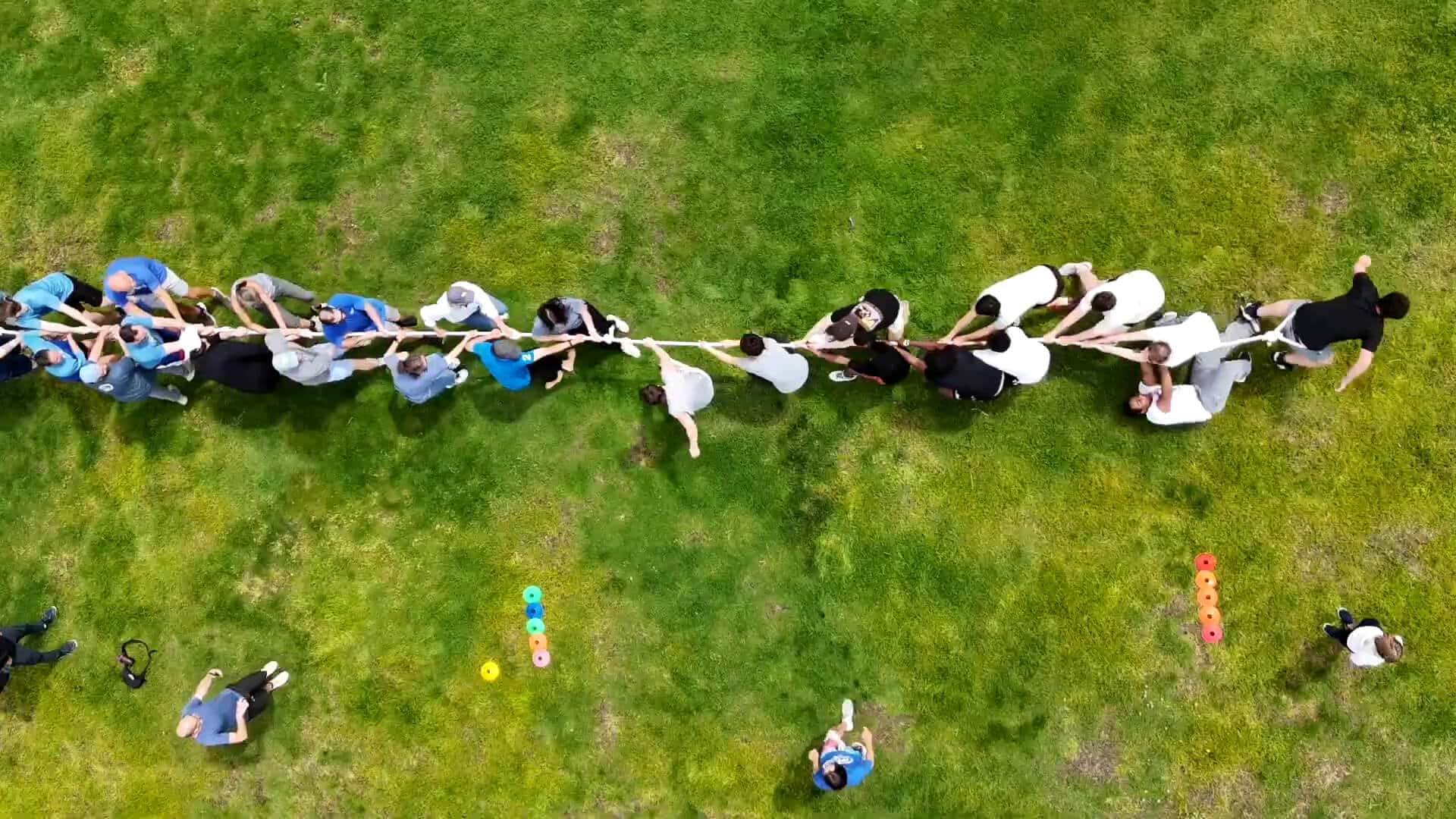 Aerial view of two teams enjoying a tug-of-war on a grassy field, capturing the spirit of a Summer Experience, with a person in blue observing nearby and colorful markers scattered on the grass.