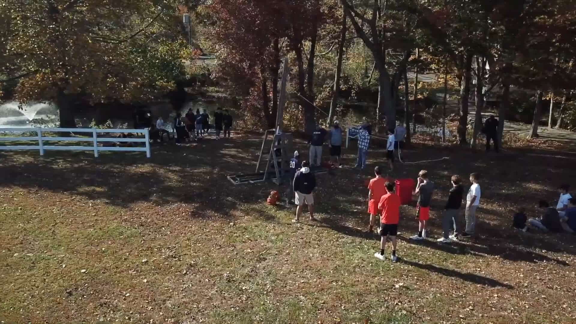 A group of people are gathered outdoors in a grassy, wooded area, enjoying a Summer Experience—some stand near a white fence and trees, while others gather by a swing set and an orange cone.