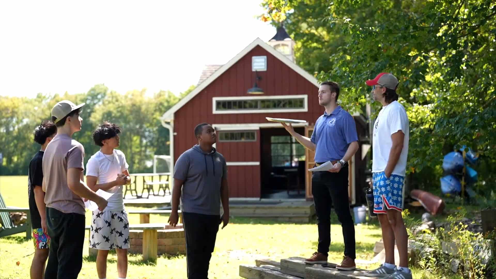 Five people stand and talk outside near a small red building; one person with a clipboard appears to be addressing the group during a Summer Experience. Trees and picnic tables are visible in the background.