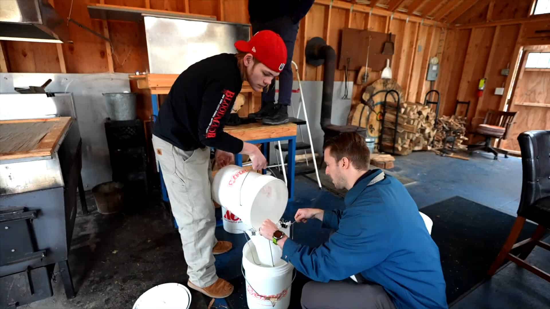 Two men enjoy a summer experience as they pour liquid from one white bucket into another inside a wooden shed, surrounded by kitchen equipment and stacked wood in the background.