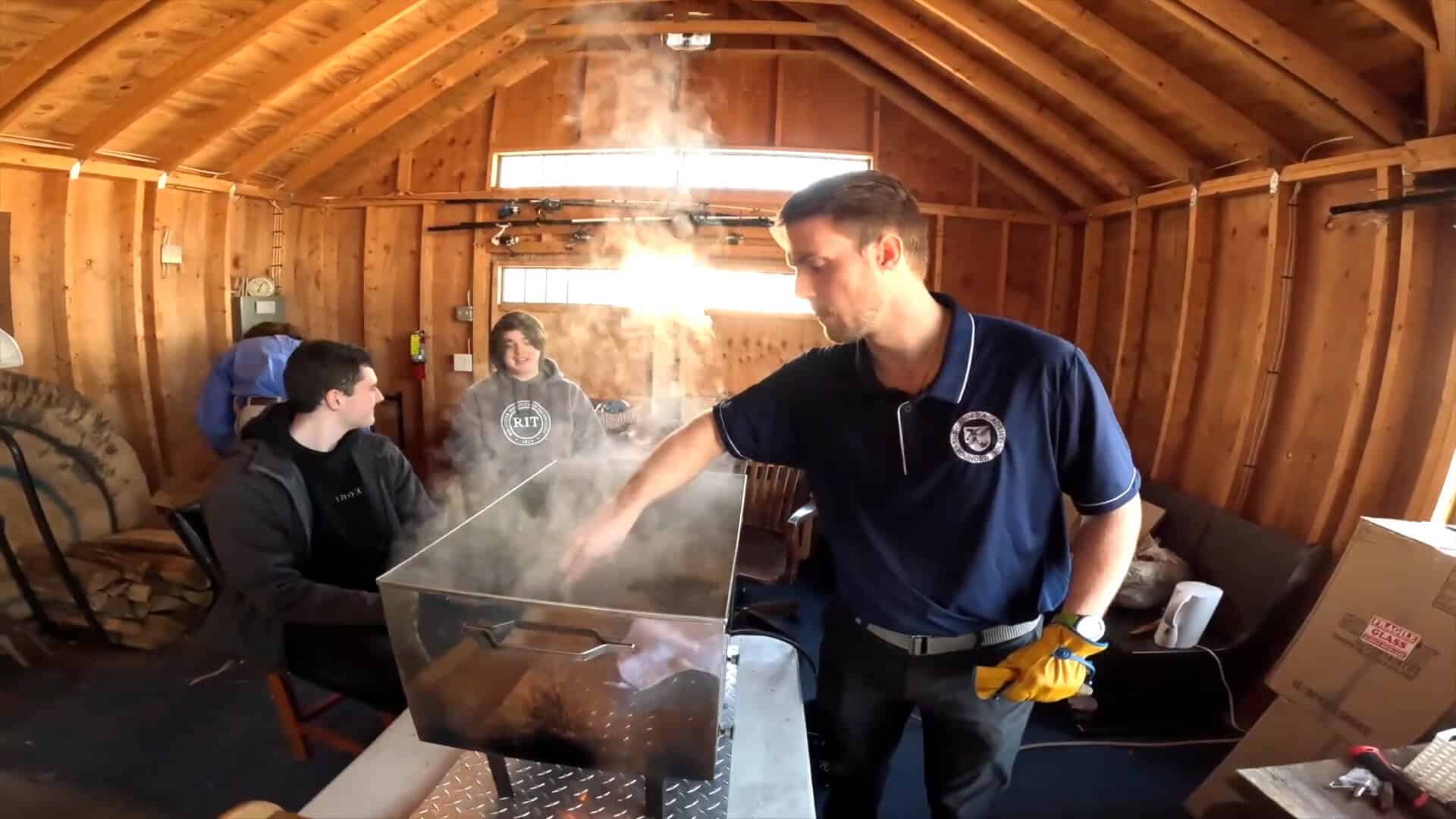 A man wearing gloves tends to a steaming metal container inside a wooden shed, while two other people sit and watch, enjoying this unique summer experience together.