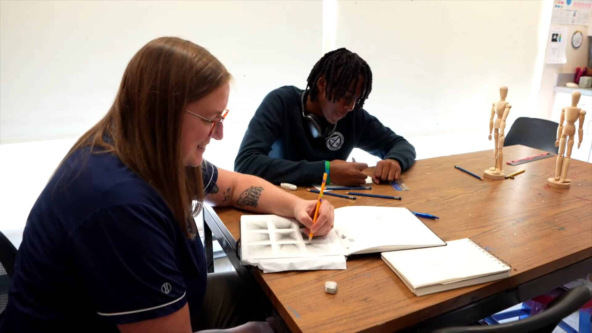 Two people sit at a table drawing in sketchbooks with pencils, surrounded by art supplies and wooden mannequins, enjoying a creative summer experience.