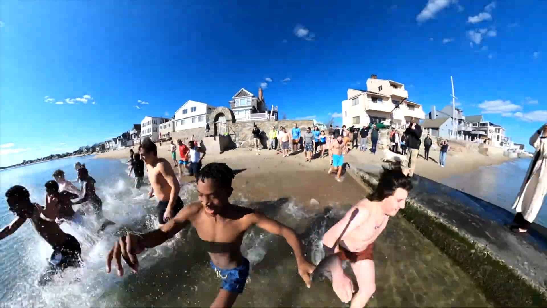 People run from the ocean onto a sandy beach, capturing the ultimate summer experience, with houses and a crowd of onlookers in the background under a clear blue sky.