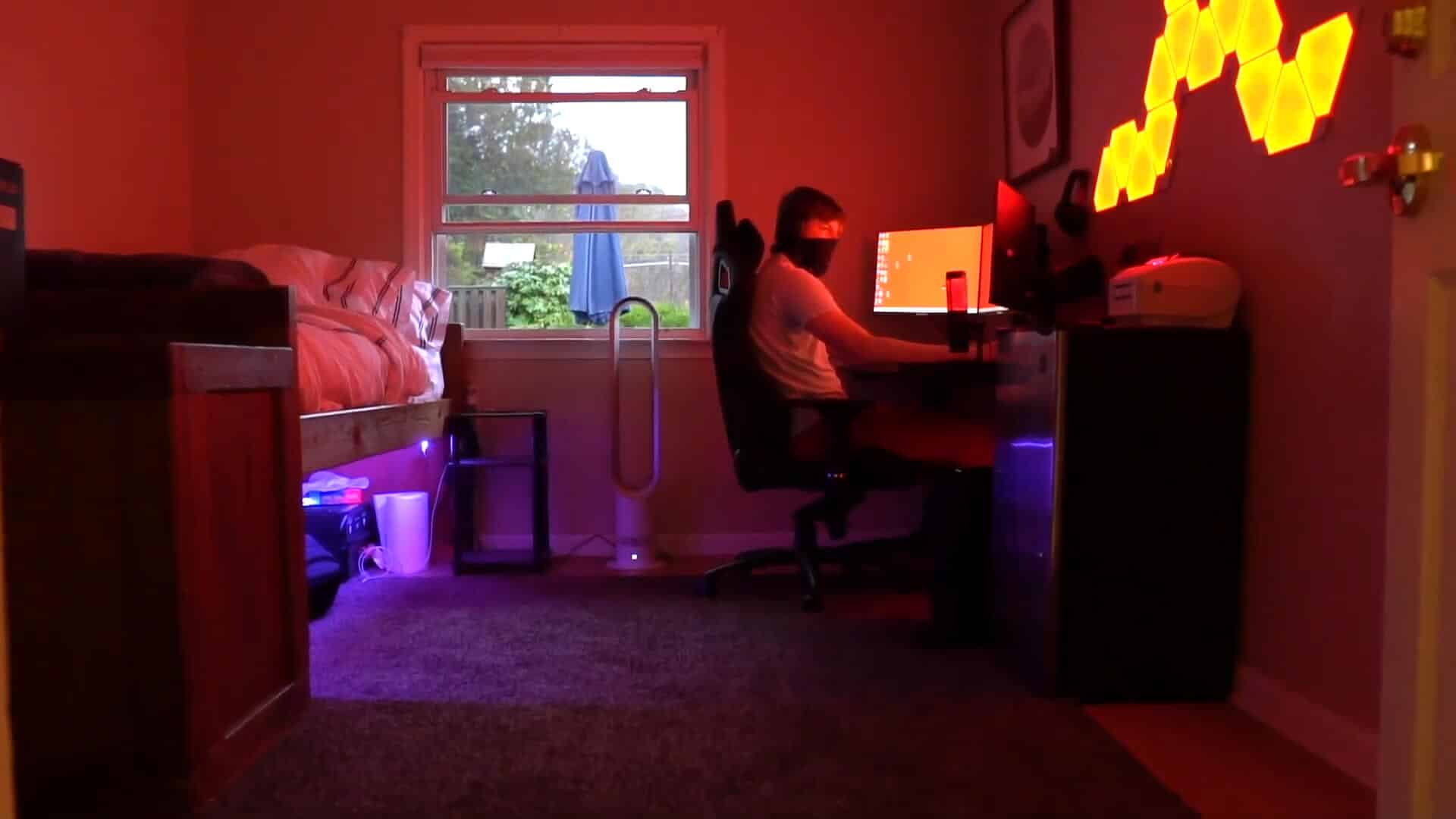 A person enjoys a summer experience, sitting at a desk using a computer in a dimly lit room with orange and purple lights, a bed on the left, and a window showing trees outside.