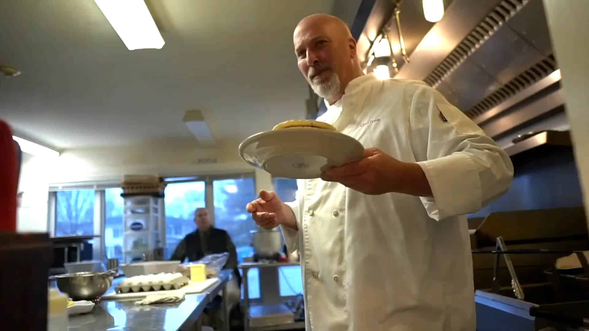 Chef in a white coat holds a plate with food in a commercial kitchen, capturing the energy of a true Summer Experience, with kitchen equipment and another person visible in the background.