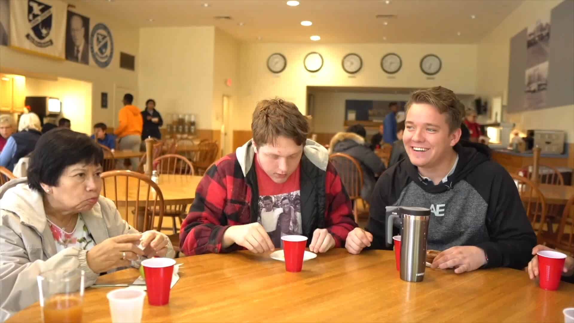 Three people enjoy their summer experience as they sit at a wooden table in a cafeteria, eating and drinking from red cups. Other people are visible in the background.
