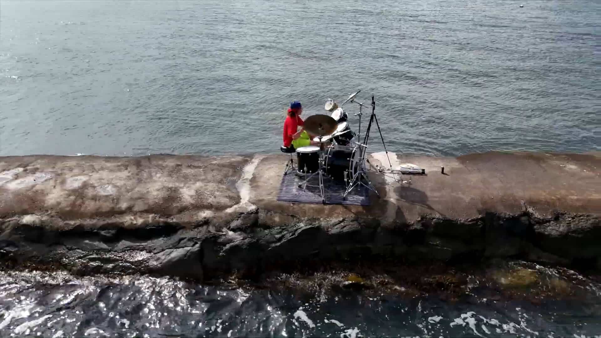 A person enjoys a unique summer experience, playing a drum set on a rocky pier by the water, with recording equipment set up nearby.