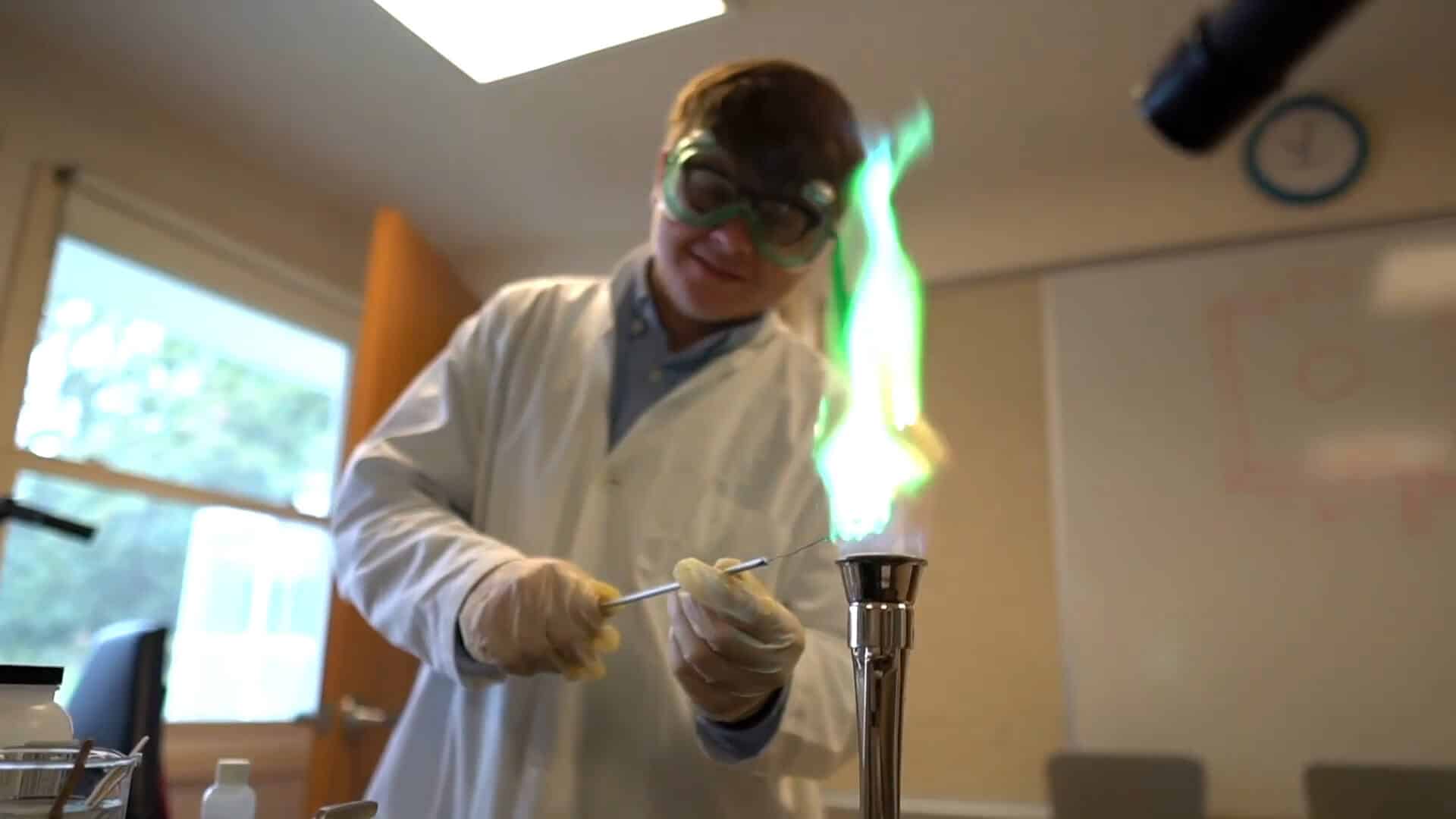 A person enjoying a Summer Experience in a science laboratory wears safety goggles and a lab coat while holding a wire over a burner with a green flame.