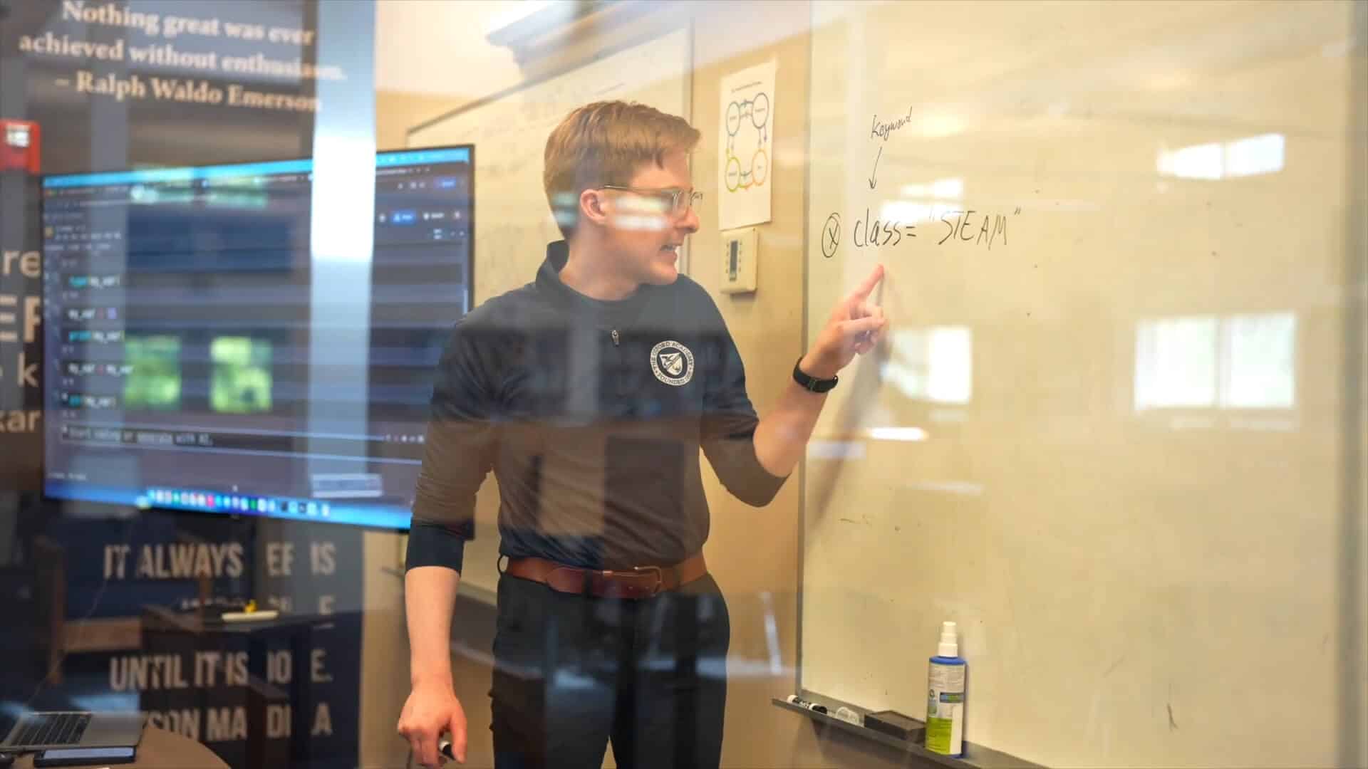 A person stands at a whiteboard, pointing to a section labeled "@class= 'DEAM'" with a marker in hand; computer screens are visible in the background, capturing the energy of a collaborative Summer Experience.