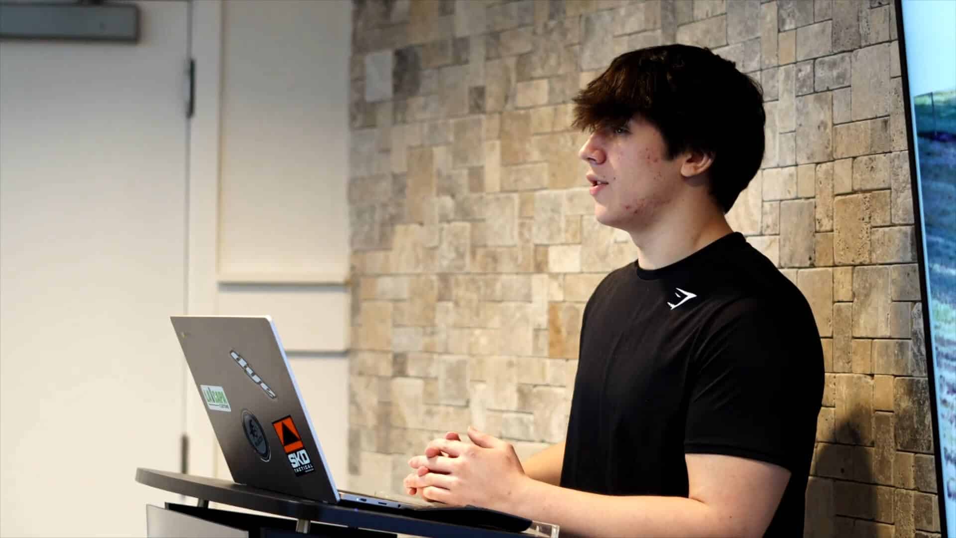 A person in a black shirt stands behind a laptop on a desk, sharing their Summer Experience while presenting in front of a stone-patterned wall.