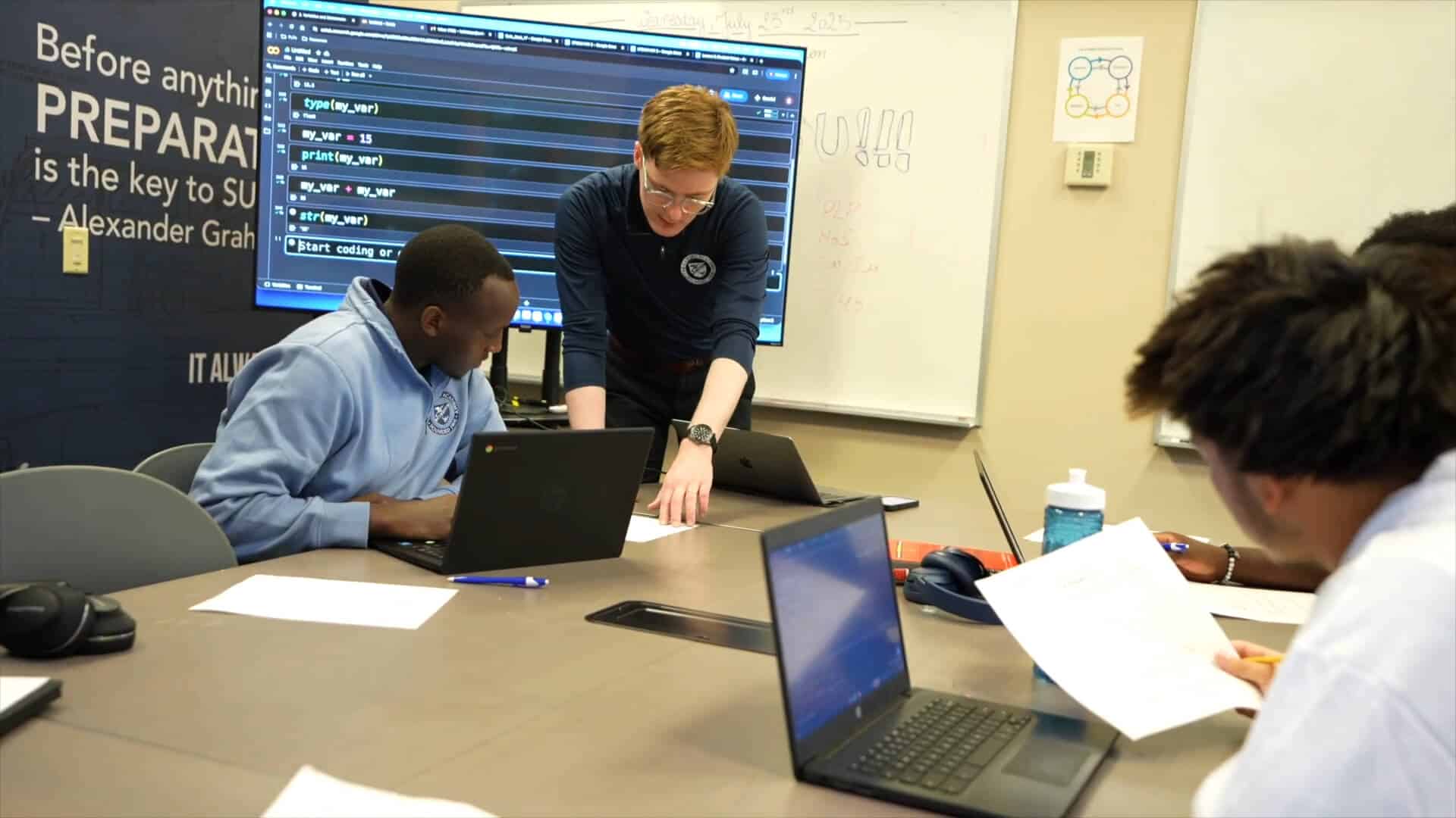 Three people sit at a conference table with laptops and papers while another stands, pointing at a document; the scene captures a collaborative Summer Experience, with a large monitor and whiteboard in the background.