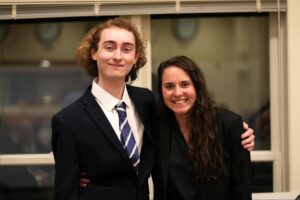 Two people in business attire stand indoors, smiling at the camera. One person has an arm around the other's shoulder. Blurred window and wall are in the background.