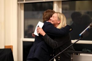Two people hug each other and smile during an indoor event, with a microphone and window in the background.