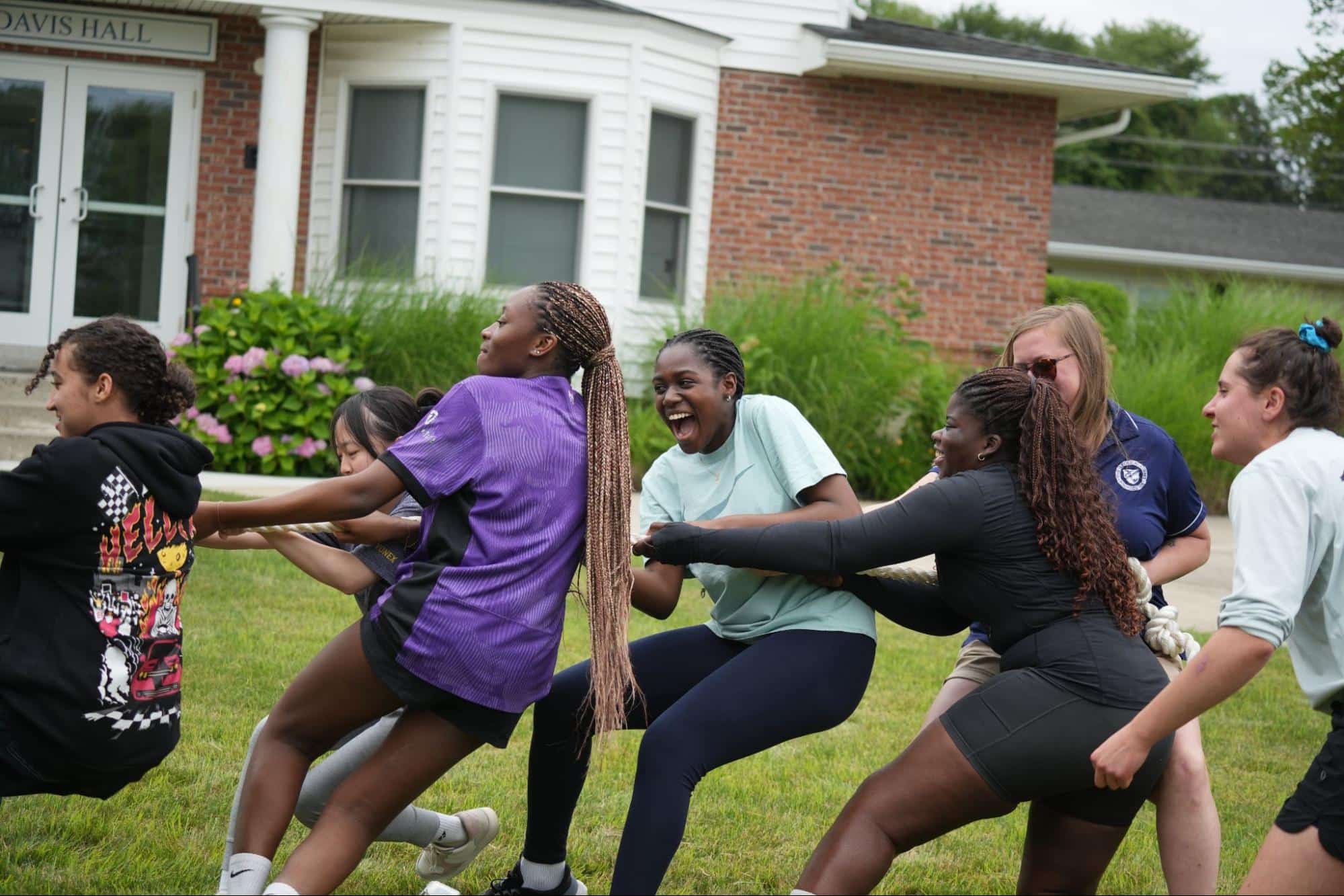 A group of people playing tug of war outside on grass in front of a brick building labeled "Davis Hall.